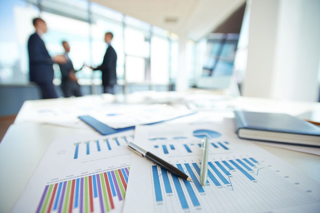Printed bar graphs and reports on a desk with a blurred background of businessmen walking by.