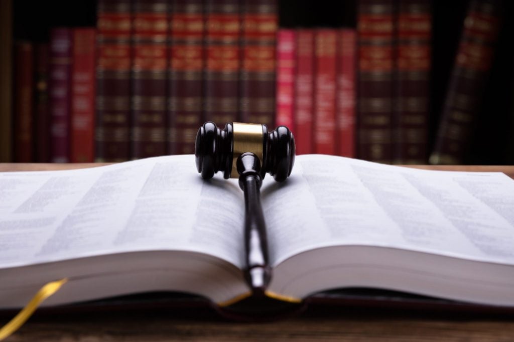 A book of law open with a gavel on top, and red books on a bookshelf behind.